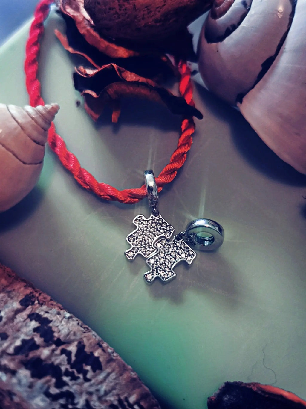 Puzzle-Anhänger aus S925-Silber mit Zirkonen auf rotem Faden, auf grünem Hintergrund mit Muscheln.