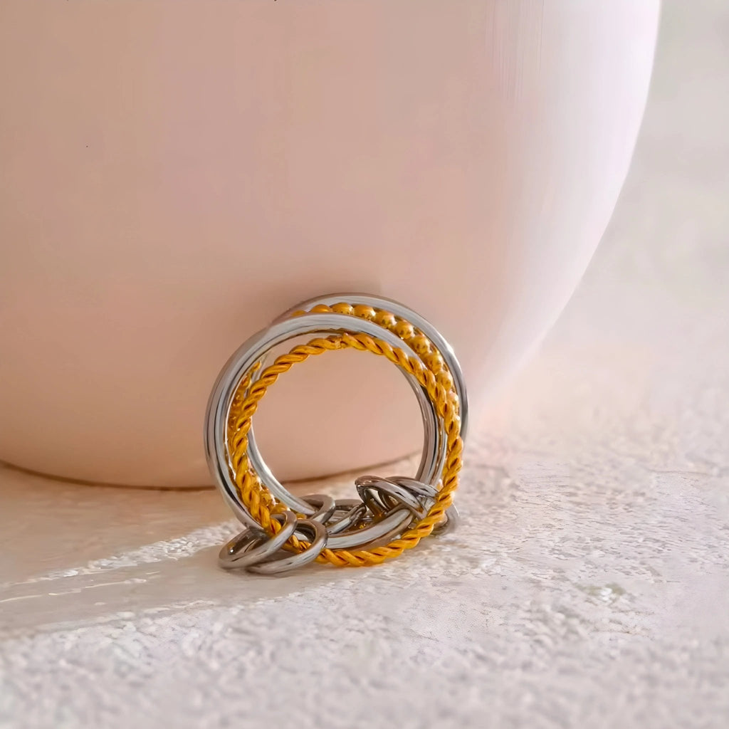 BEHI MILANO Onda zweifarbiger Ring mit drei Bändern: zwei aus poliertem Stahl und eines ineinander verschlungen, 18 Karat vergoldet, auf hellem Hintergrund.