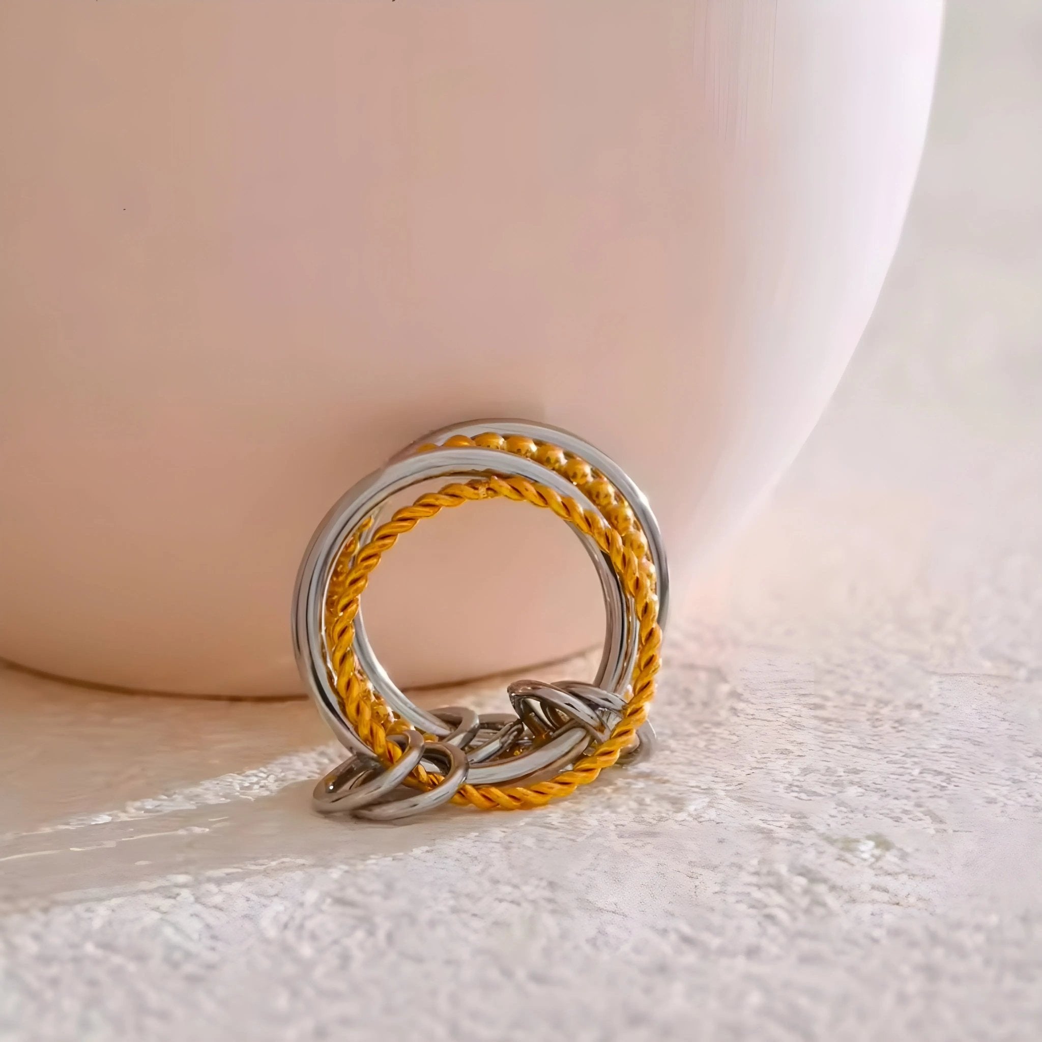 Anello BEHI MILANO Onda bicolore con tre fasce: due in acciaio lucido e una placcata oro 18k intrecciata, su sfondo chiaro.