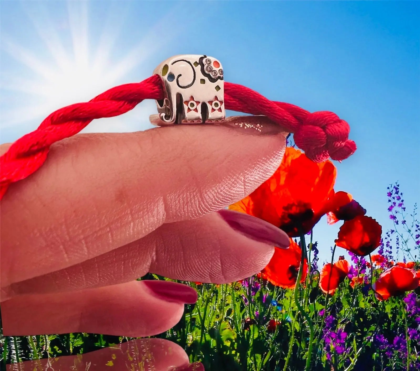 Charm Ganesh elefante argento S925 decorato su filo rosso indossato su un dito con fiori rossi e cielo azzurro