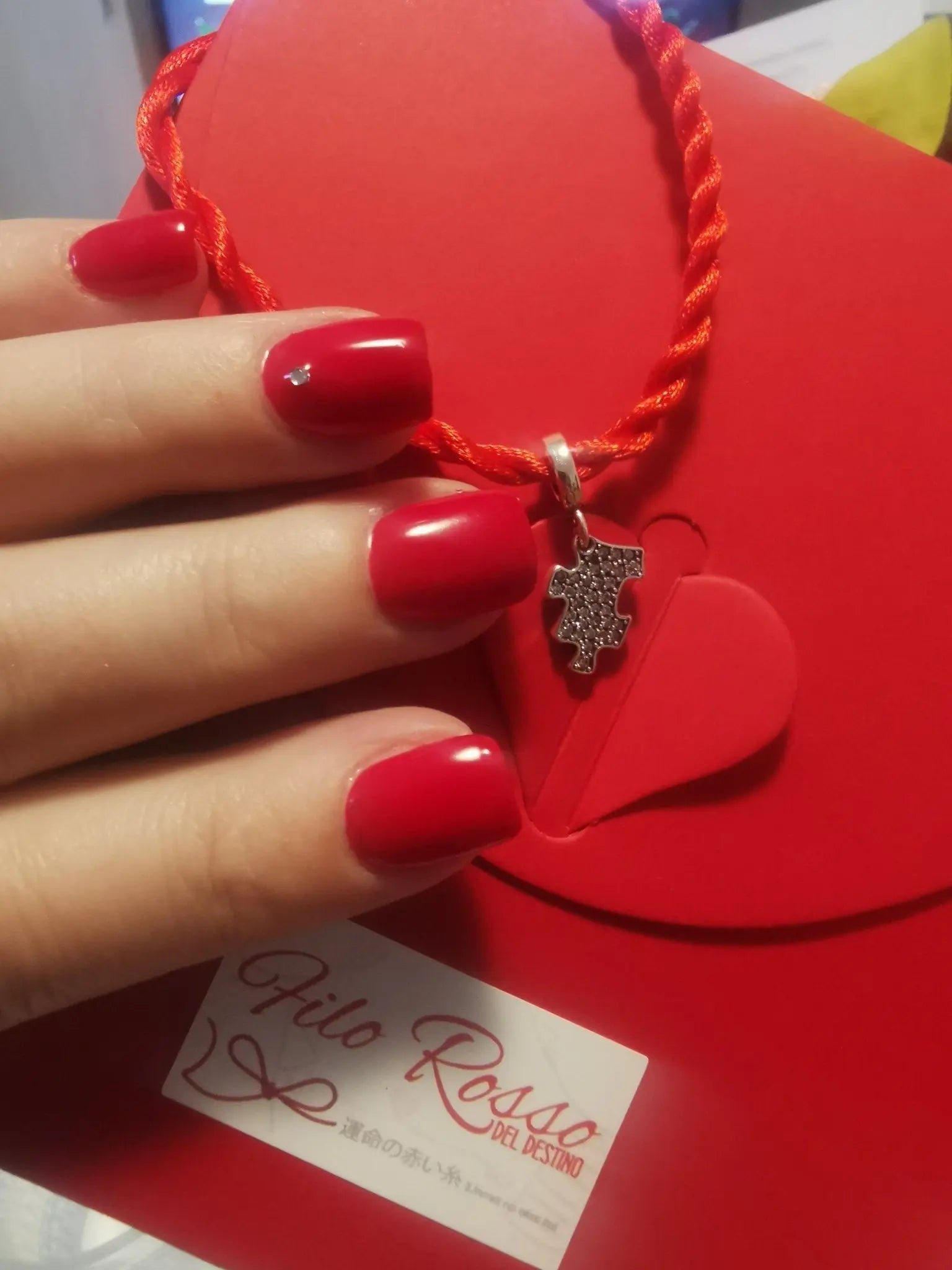 Mano con unghie rosse tiene bracciale Filo Rosso con charm puzzle argento S925, su confezione regalo rossa.