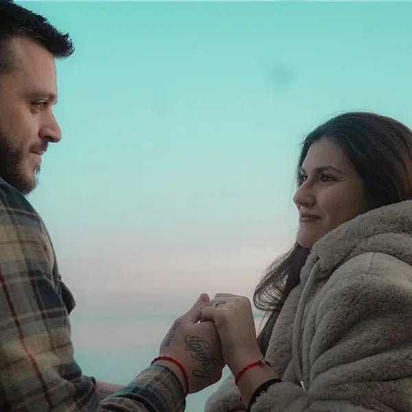 Coppia che si tiene per mano, entrambi con braccialetti Filo Rosso e Charm Hamsa argento al polso, sotto cielo azzurro.